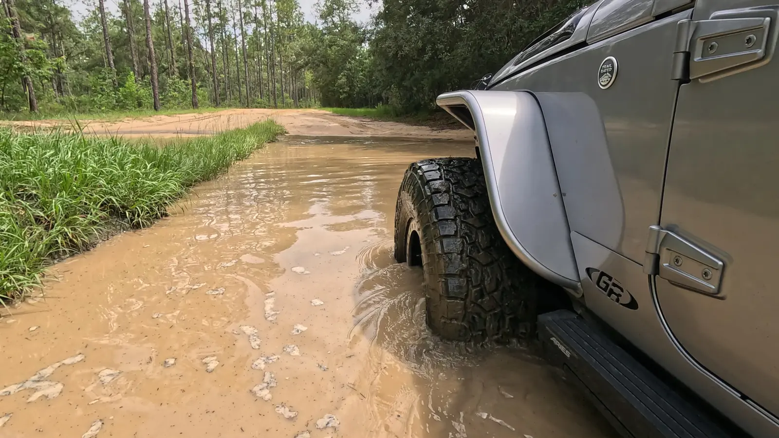 Silver Jeep Wrangler crossing a muddy water hole on a Florida pine-flatwoods trail